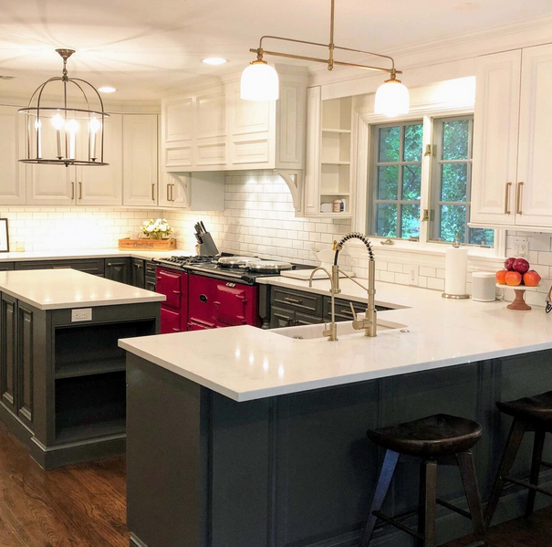 This image shows a modern kitchen with white upper cabinets, a dark gray island with white countertops, a red range, subway tile backsplash, and pendant lighting fixtures.