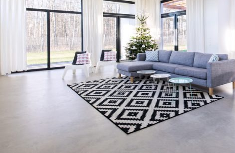 This image shows a modern minimalist living room with a gray sectional sofa, black and white geometric patterned rug, large black-framed windows, and polished concrete floors.