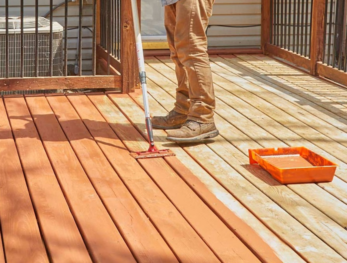 This image shows a person staining or sealing a wooden deck with a roller, demonstrating the contrast between the treated darker wood planks and the untreated lighter ones, with an orange paint tray visible nearby.