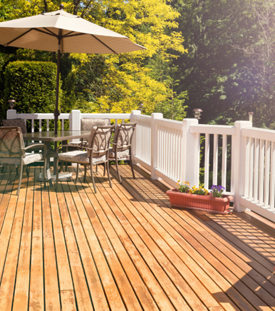 This image shows a beautiful wooden deck with white railings, outdoor furniture including chairs and a table with an umbrella, and planters with flowers, all surrounded by lush green trees in bright sunlight.