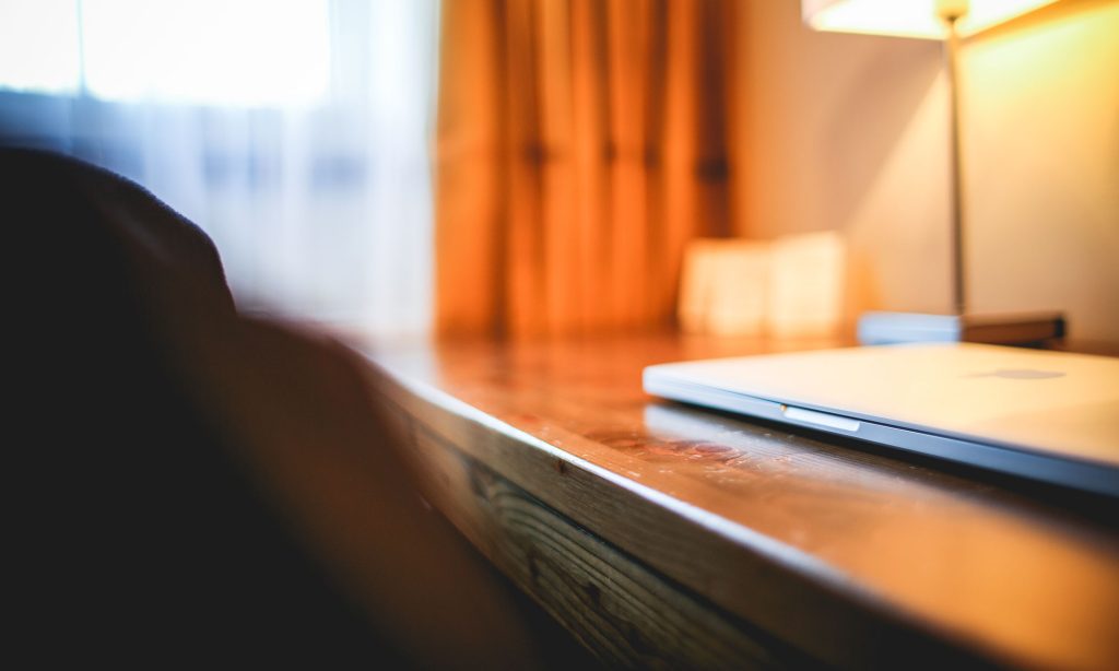 This image shows a close-up view of a wooden desk or table surface with a laptop and what appears to be a smartphone, with warm orange curtains and soft lighting visible in the blurred background.