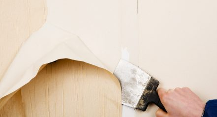 The image shows a person using a putty knife to peel off wallpaper from a wall as part of a wallpaper removal process.
